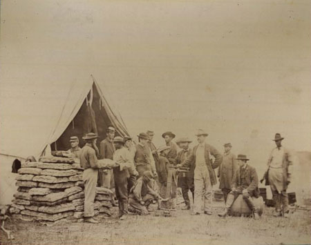 The photograph shows the Commissary Department at the Headquarters of the Army of the Potomac in Fairfax Courthouse, VA. The gentleman with his hand on the scale is Captain John R. Coxe with his staff. The large stacks of freshly baked loaves of soft bread were prepared for issuance to the troops.