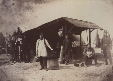 This photograph shows a different version of a cook house during camp in Hilton Head in 1862. Pictured from left to right is J.N. Campbell, Corporal McQueston, General Wheeler, A.H. Stevens, W. Blake, and Captain W.H. Maxwell of Company H of the 3rd New Hampshire Infantry. The Company cooks are wearing the aprons, while the other gentlemen are most likely waiting for their food or coffee to be ready.